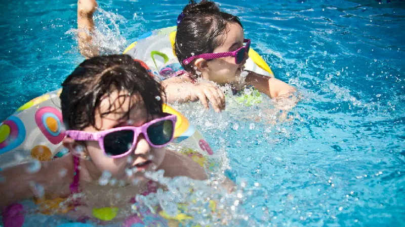 Two young girls in the pool wearing ring floats and sunglasses and splashing around having fun.